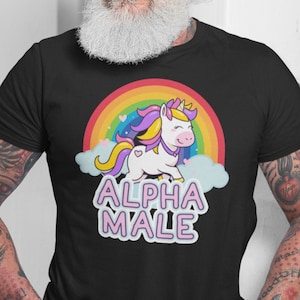 May include: Black t-shirt featuring a cartoon unicorn with a rainbow and clouds. The unicorn has a yellow horn, purple and yellow mane, and is set against a rainbow. The text "ALPHA MALE" is below the unicorn in a pink and white font.