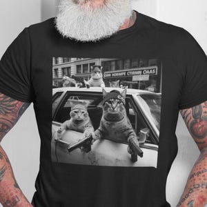 May include: Black t-shirt featuring a black and white image of cats in a car, with one cat holding a toy gun. The image has a vintage, film-like quality. The cats are wearing bandanas and hats. The shirt is worn by a person with a white beard.