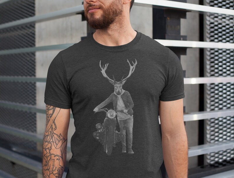 May include: Dark gray t-shirt featuring a graphic of a deer-headed figure in a jacket and jeans, leaning on a vintage motorcycle. The design is in black and white. The shirt is a crew neck.