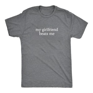 May include: A heather gray t-shirt with the text "my girlfriend beats me" in white, centered on the chest. The shirt has short sleeves and a crew neck.