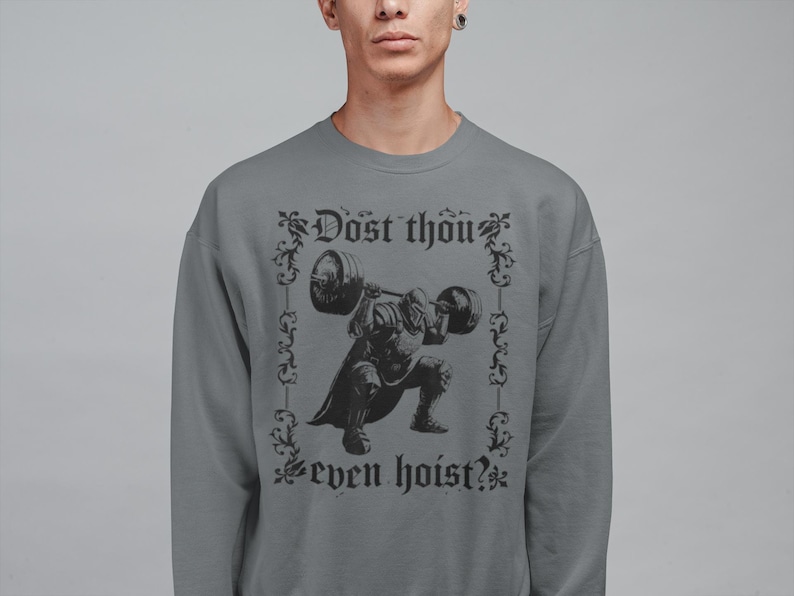 Op de afbeelding: Grijze sweatshirt met een zwarte afbeelding van een middeleeuwse ridder die een halter optilt. De tekst "Dost thou even hoist?" is boven en onder de ridderillustratie in een decoratief lettertype gedrukt. Ronde hals.