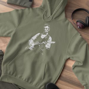 Peut inclure: Un sweat &agrave; capuche vert olive avec un motif blanc d'Abraham Lincoln jouant de la guitare &eacute;lectrique. L'image comprend &eacute;galement un bonnet assorti, des baskets noires et un casque avec des oreillettes marron.