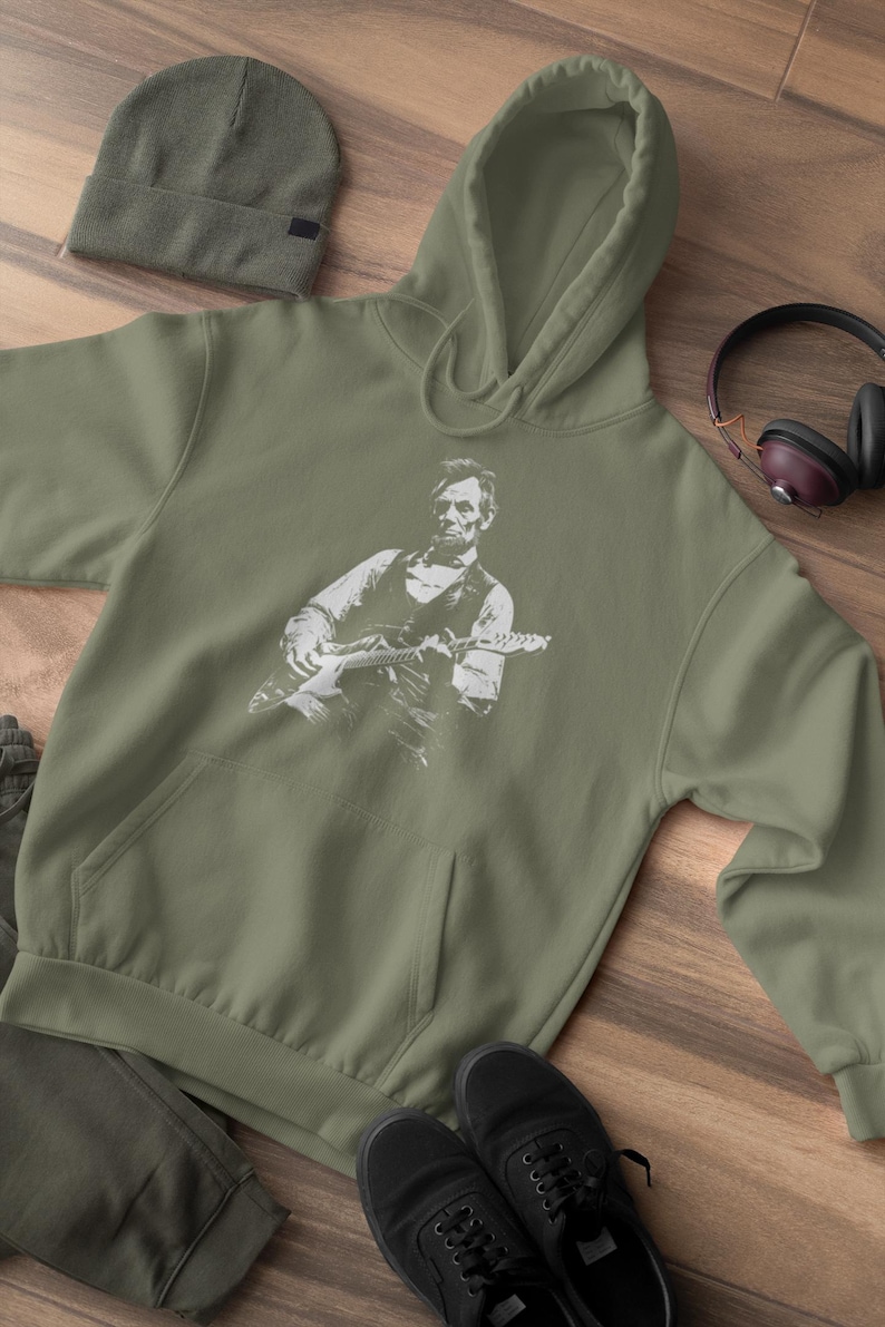 Peut inclure: Un sweat &agrave; capuche vert olive avec un motif blanc d'Abraham Lincoln jouant de la guitare &eacute;lectrique. L'image comprend &eacute;galement un bonnet assorti, des baskets noires et un casque avec des oreillettes marron.