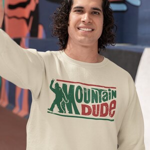May include: A cream-colored sweatshirt with a green and red graphic of a person with the text "Mountain Dude".
