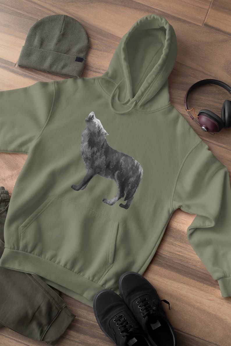 May include: A green hooded sweatshirt with a black and white wolf howling graphic print. The sweatshirt is laid out on a wooden surface with a beanie, headphones, and shoes.
