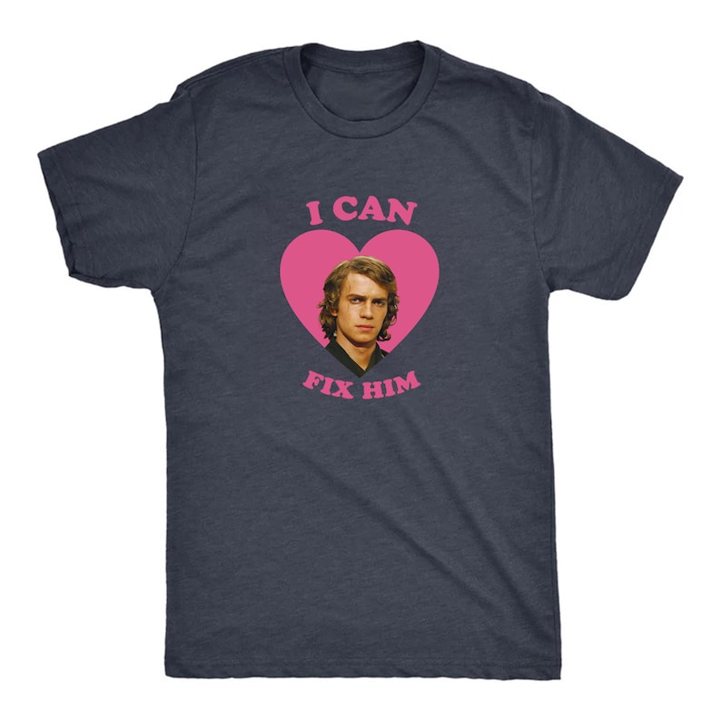 May include: Dark navy blue t-shirt with a pink heart graphic. The heart contains a portrait of a man and the text "I CAN FIX HIM" in pink. The shirt is a crew neck.