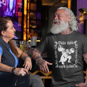 Op de afbeelding: Een zwart t-shirt met de tekst "Dost thou even hoist?" in het wit, met een gewichtheffer grafiek. Het shirt wordt gedragen door een persoon met een lange witte baard en tatoeages. Een vrouw zit naast de persoon.