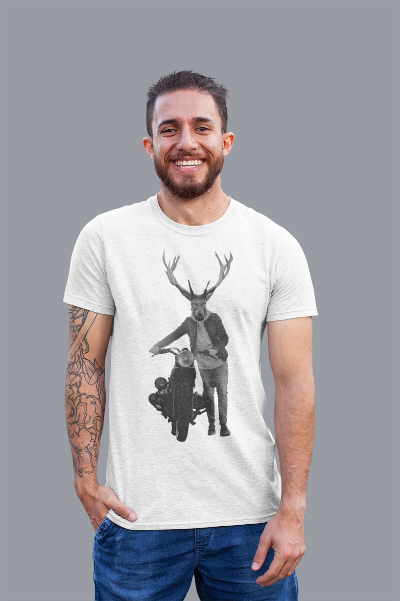 May include: A light gray t-shirt featuring a black and white graphic of a deer-headed figure standing next to a vintage motorcycle. The figure is wearing a jacket and jeans. The person wearing the shirt is smiling.