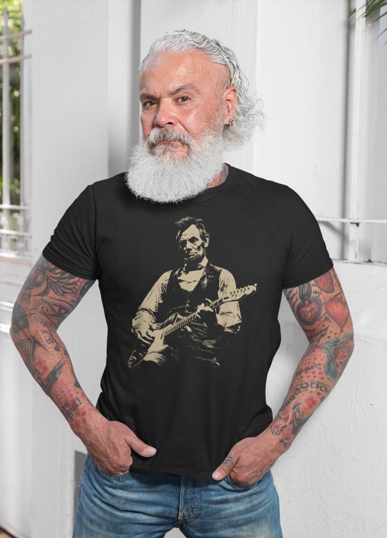 Peut inclure: T-shirt noir avec une image s&eacute;pia d'Abraham Lincoln jouant de la guitare. L'image montre Lincoln en gilet et chemise, tenant la guitare. La personne portant le t-shirt a des tatouages sur les bras.