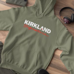Puede incluir: Una sudadera con capucha verde oliva con el texto "KIRKLAND Signature" en blanco y rojo. La sudadera tiene un bolsillo delantero y una capucha. Tambi&eacute;n se ven un gorro a juego, zapatos negros y auriculares.