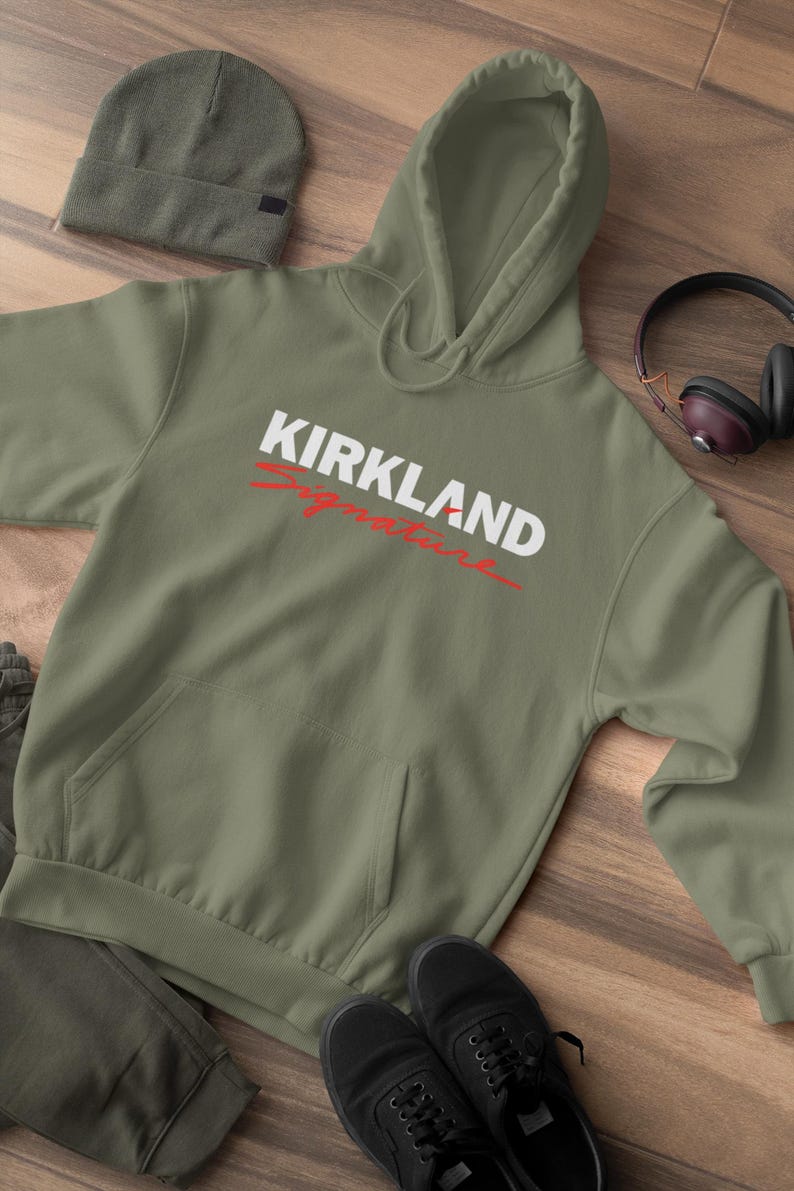 Puede incluir: Una sudadera con capucha verde oliva con el texto "KIRKLAND Signature" en blanco y rojo. La sudadera tiene un bolsillo delantero y una capucha. Tambi&eacute;n se ven un gorro a juego, zapatos negros y auriculares.