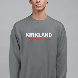 Puede incluir: Sudadera gris de cuello redondo con el texto "KIRKLAND Signature" en blanco y rojo. La sudadera es de color liso y tiene un dise&ntilde;o sencillo y cl&aacute;sico.