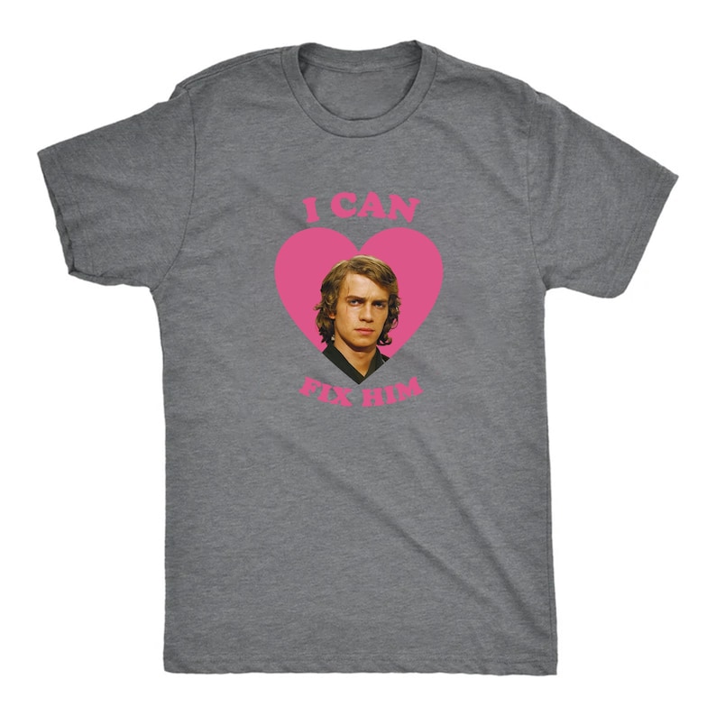 May include: A heather gray t-shirt featuring a pink heart with the image of a man inside. The text "I CAN FIX HIM" is printed above and below the heart in pink. The shirt is a crew neck style.