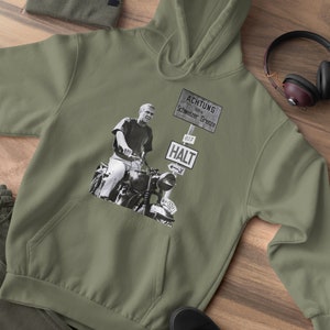 May include: A green hooded sweatshirt with a black and white image of a man riding a motorcycle. The image includes a sign that reads "ACHTUNG 100m Schweizer Grenze 607 HALT".