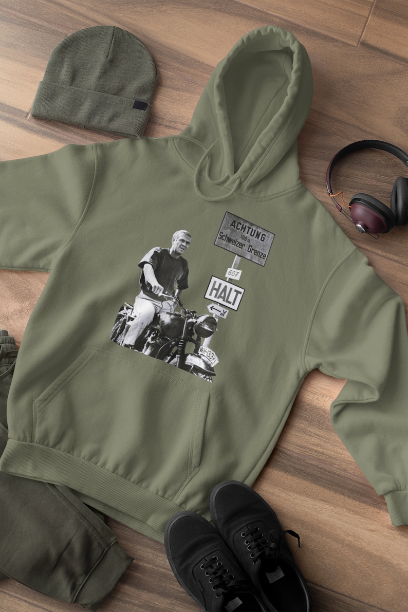 May include: A green hooded sweatshirt with a black and white image of a man riding a motorcycle. The image includes a sign that reads "ACHTUNG 100m Schweizer Grenze 607 HALT".