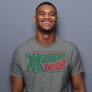 Może przedstawiać: Szary t-shirt z nadrukiem w kolorze zielonym i czerwonym przedstawiającym postać z kresk&oacute;wki trzymającą puszkę Mountain Dew i napis "Mountain Dude".