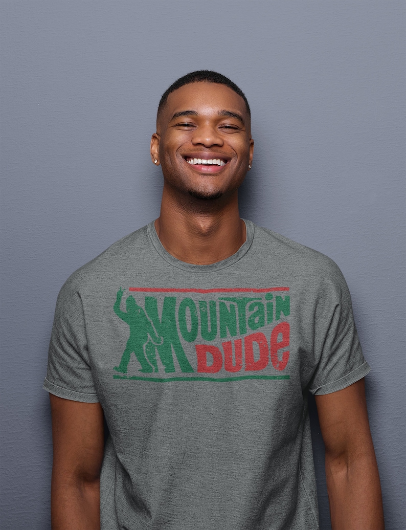 Może przedstawiać: Szary t-shirt z nadrukiem w kolorze zielonym i czerwonym przedstawiającym postać z kresk&oacute;wki trzymającą puszkę Mountain Dew i napis "Mountain Dude".