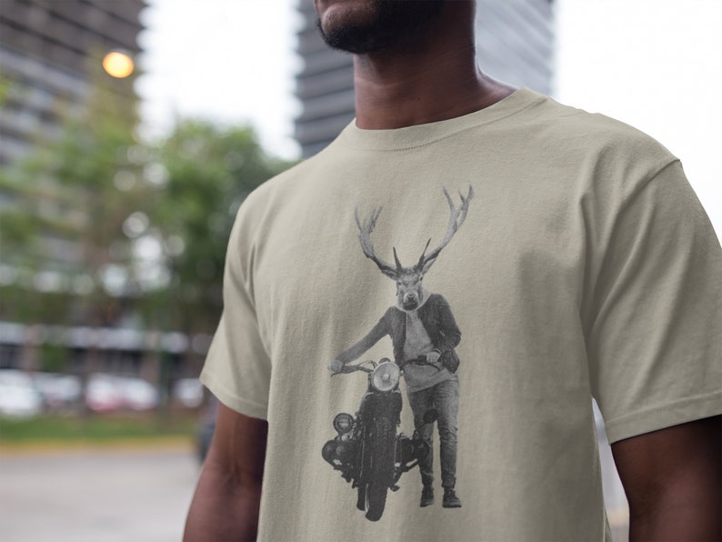May include: Beige t-shirt featuring a black and white illustration of a deer-headed figure leaning on a vintage motorcycle. The figure is wearing a jacket, jeans, and boots. The motorcycle has a round headlight and a classic design.