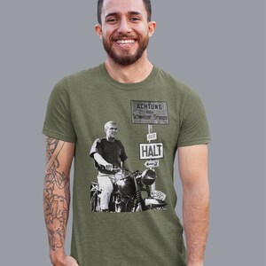 May include: A green t-shirt with a black and white image of a man on a motorcycle. The image is of a sign that reads "ACHTUNG 100 m Schweizer Grenze 607 HALT" with an arrow pointing left. The motorcycle has the license plate "NH 13371".