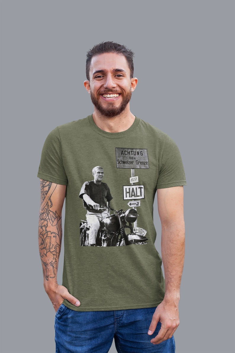 May include: A green t-shirt with a black and white image of a man on a motorcycle. The image is of a sign that reads "ACHTUNG 100 m Schweizer Grenze 607 HALT" with an arrow pointing left. The motorcycle has the license plate "NH 13371".