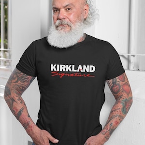 Puede incluir: Camiseta negra de cuello redondo con el texto "KIRKLAND Signature" en blanco y rojo. La camiseta la lleva una persona con barba blanca y tatuajes en los brazos.