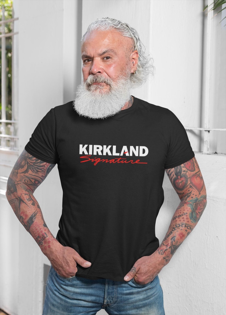 Puede incluir: Camiseta negra de cuello redondo con el texto "KIRKLAND Signature" en blanco y rojo. La camiseta la lleva una persona con barba blanca y tatuajes en los brazos.