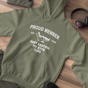 May include: Olive green hoodie with the text "PROUD MEMBER OF THE JUST RESTING MY EYES CLUB" in white. A matching beanie, black shoes, and headphones are also visible. The hoodie has a front pocket and a drawstring hood.
