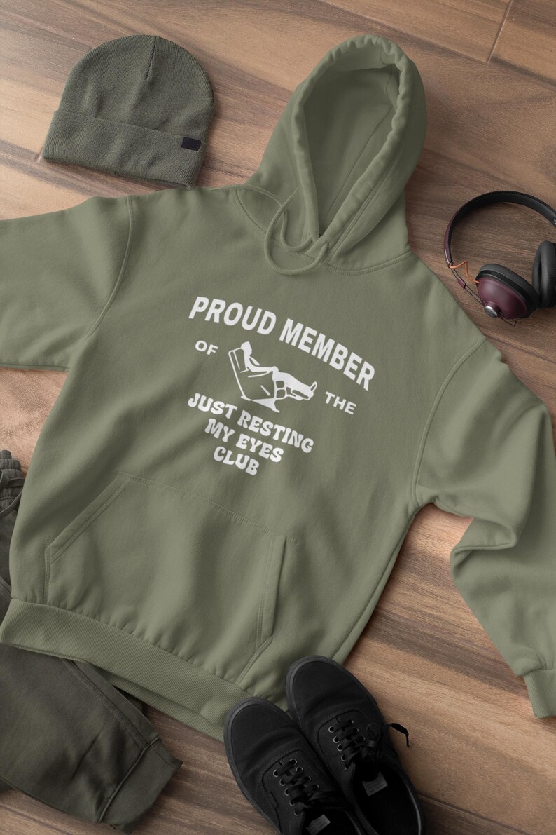 May include: Olive green hoodie with the text "PROUD MEMBER OF THE JUST RESTING MY EYES CLUB" in white. A matching beanie, black shoes, and headphones are also visible. The hoodie has a front pocket and a drawstring hood.
