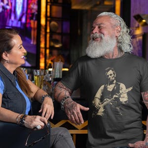 Peut inclure: Un t-shirt noir avec une illustration d'Abraham Lincoln jouant de la guitare. Un homme et une femme sont assis &agrave; un bar. La femme porte une chemise bleue et un gilet en jean. L'homme porte le t-shirt et a des tatouages sur les bras.