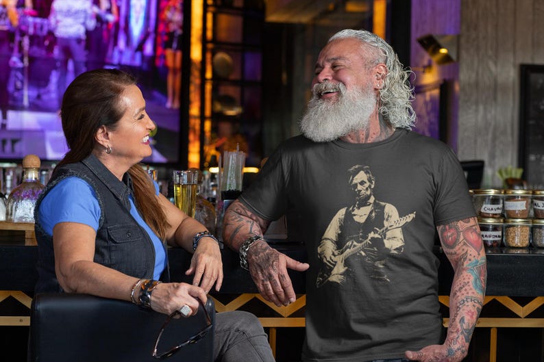 Peut inclure: Un t-shirt noir avec une illustration d'Abraham Lincoln jouant de la guitare. Un homme et une femme sont assis &agrave; un bar. La femme porte une chemise bleue et un gilet en jean. L'homme porte le t-shirt et a des tatouages sur les bras.