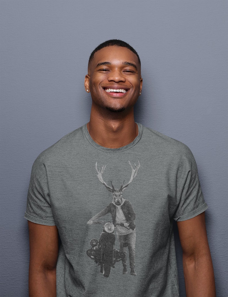 May include: Gray t-shirt featuring a graphic of a deer-headed figure in a jacket and jeans, leaning on a vintage motorcycle. The design is in black and white. The person wearing the shirt is smiling.