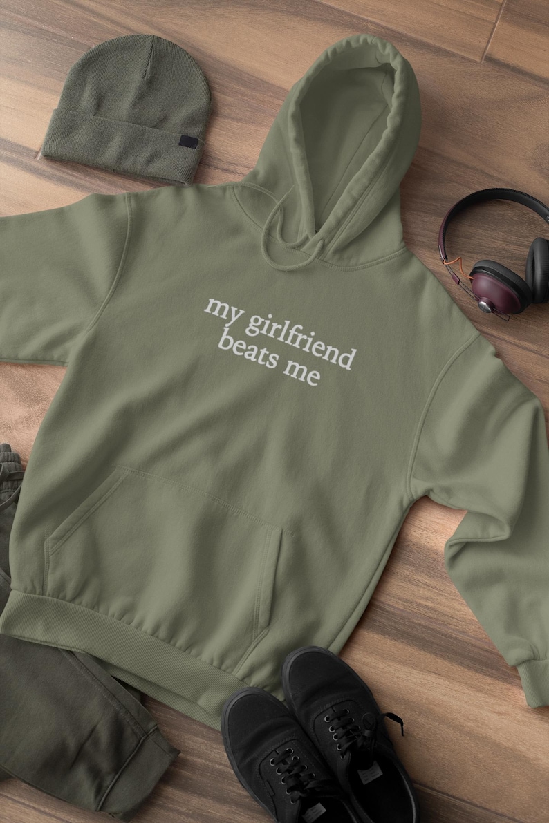 May include: Olive green hoodie with the text "my girlfriend beats me" in white. The hoodie has a front pocket and a drawstring hood. A matching beanie, black shoes, and headphones are also visible.