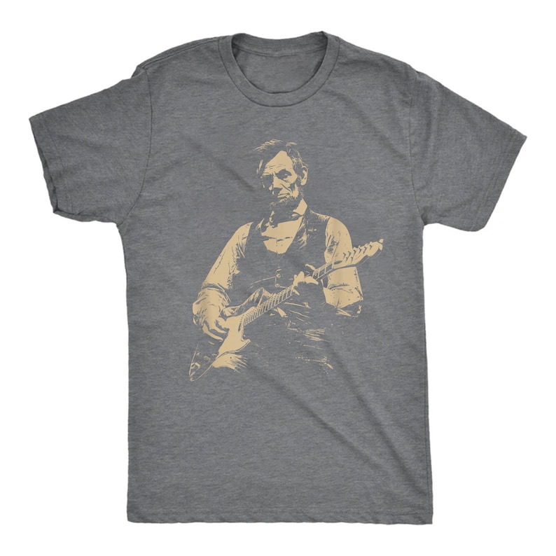 Peut inclure: T-shirt gris avec une image s&eacute;pia d'Abraham Lincoln jouant de la guitare &eacute;lectrique. Le graphisme montre Lincoln en gilet et chemise, tenant la guitare. Le t-shirt est fait d'une mati&egrave;re douce.
