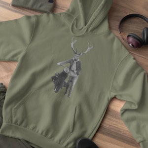 May include: Olive green hoodie with a kangaroo pocket, featuring a black and white graphic of a deer-headed figure with a motorcycle. Includes a matching beanie, sweatpants, and black sneakers. Headphones are visible.