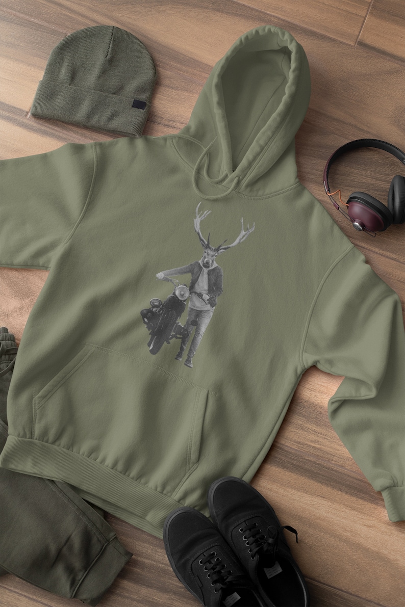 May include: Olive green hoodie with a kangaroo pocket, featuring a black and white graphic of a deer-headed figure with a motorcycle. Includes a matching beanie, sweatpants, and black sneakers. Headphones are visible.
