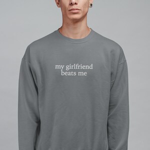 May include: A gray sweatshirt with the text "my girlfriend beats me" in white. The crew neck sweatshirt is a solid color and has long sleeves. The text is centered on the chest.