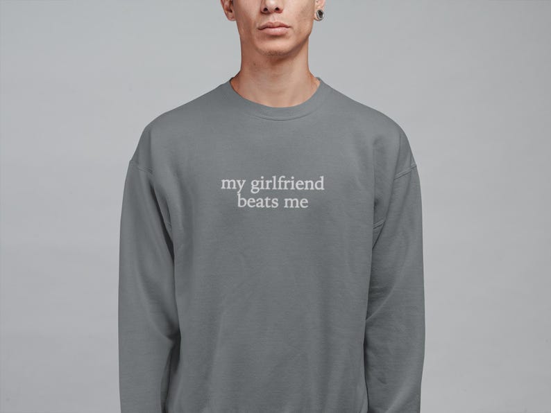 May include: A gray sweatshirt with the text "my girlfriend beats me" in white. The crew neck sweatshirt is a solid color and has long sleeves. The text is centered on the chest.