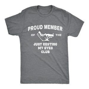 May include: A heather gray t-shirt with white text that reads "PROUD MEMBER OF THE JUST RESTING MY EYES CLUB." The shirt features a graphic of a hand with closed eyes. The t-shirt has short sleeves and a crew neck.
