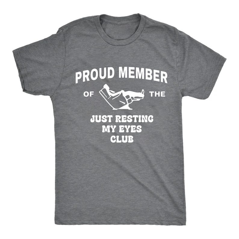 May include: A heather gray t-shirt with white text that reads "PROUD MEMBER OF THE JUST RESTING MY EYES CLUB." The shirt features a graphic of a hand with closed eyes. The t-shirt has short sleeves and a crew neck.