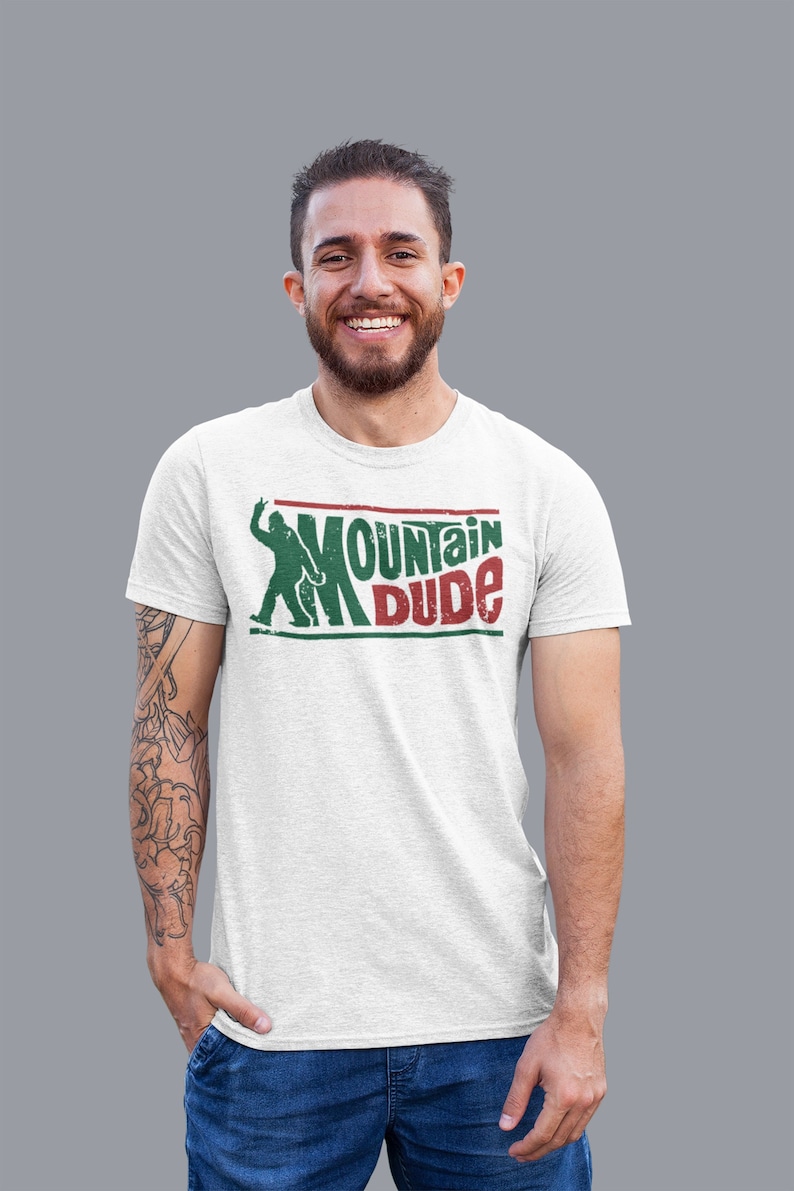 Może przedstawiać: Biała koszulka z zielonym i czerwonym nadrukiem, na kt&oacute;rym widnieje napis "Mountain Dude" oraz sylwetka osoby wędrującej.