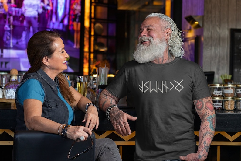May include: A man with a long white beard and tattoos is wearing a black t-shirt with white text that reads "ISWYDT". He is smiling and looking to the right. A woman with long brown hair is sitting to the left of him.
