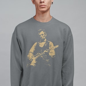 Peut inclure: Un sweat-shirt gris avec une illustration dor&eacute;e d'une personne jouant de la guitare &eacute;lectrique. L'illustration repr&eacute;sente une personne en gilet et chemise. Le sweat-shirt est de couleur unie et a un col rond.