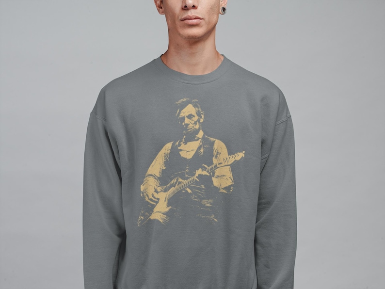 Peut inclure: Un sweat-shirt gris avec une illustration dor&eacute;e d'une personne jouant de la guitare &eacute;lectrique. L'illustration repr&eacute;sente une personne en gilet et chemise. Le sweat-shirt est de couleur unie et a un col rond.