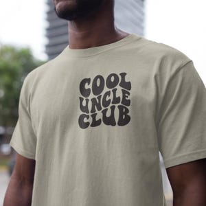 Könnte beinhalten: Ein hellbeiges T-Shirt mit einem schwarzen Grafikdruck, auf dem in einer Retro-Schrift "COOL UNCLE CLUB" steht.