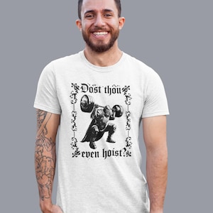 Op de afbeelding: Wit T-shirt met een zwart-wit illustratie van een gepantserde gewichtheffer die een halter optilt. De tekst "Dost thou even hoist?" staat in een decoratief kader. Het T-shirt wordt door een persoon gedragen.