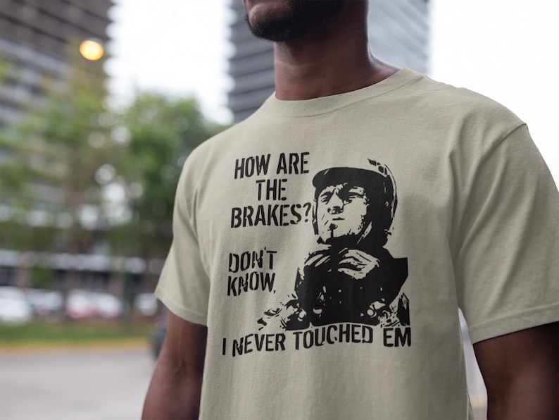 May include: A beige t-shirt with a black graphic print of a man in a helmet. The text on the shirt reads "How are the brakes? Don't know. I never touched 'em."