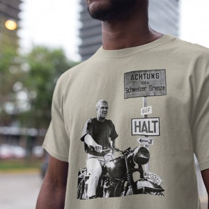 May include: A beige t-shirt with a black and white image of a man on a motorcycle. The image includes a sign that reads "ACHTUNG 100 m Schweizer Grenze" and another sign that reads "HALT 607".