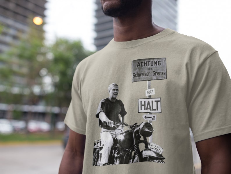 May include: A beige t-shirt with a black and white image of a man on a motorcycle. The image includes a sign that reads "ACHTUNG 100 m Schweizer Grenze" and another sign that reads "HALT 607".