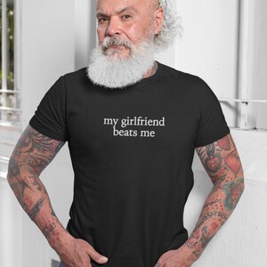 May include: Black t-shirt with the text "my girlfriend beats me" in white. The shirt is worn by a person with a white beard and tattoos on their arms. The person is wearing blue jeans.
