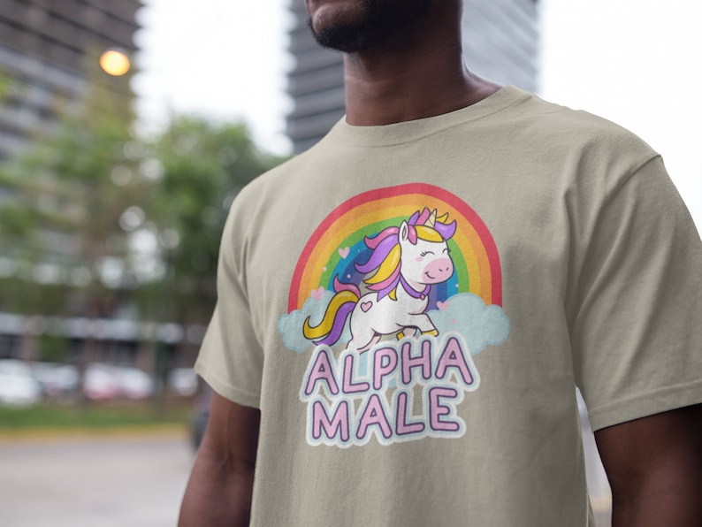 May include: A beige t-shirt featuring a cartoon unicorn with a rainbow and the text "ALPHA MALE" in pink and purple. The unicorn has a yellow horn, purple and yellow hair, and a pink heart on its side. The shirt is made of a soft material.